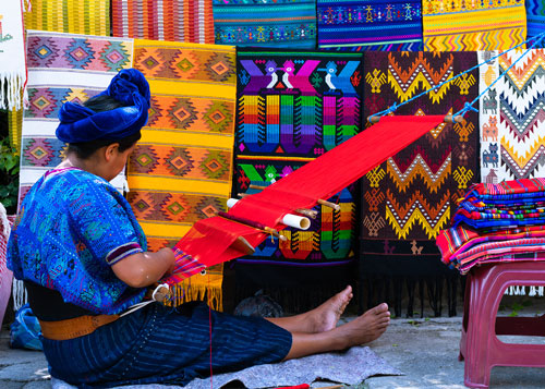 A woman weaving red threads in a loom with colorful blankets behind her