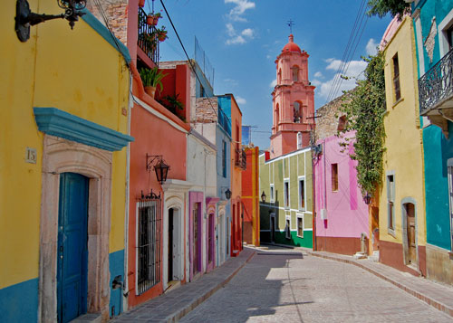 Colorful adobe and cement buildings line a street in mexico with an orange tower at the end of the road
